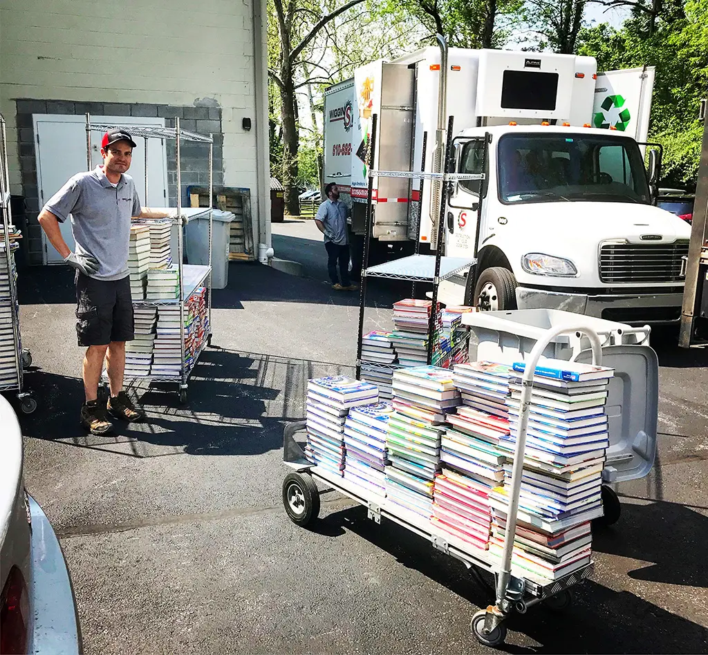 A man standing with multiple large carts of textbooks beside a Wiggins Shredding Truck