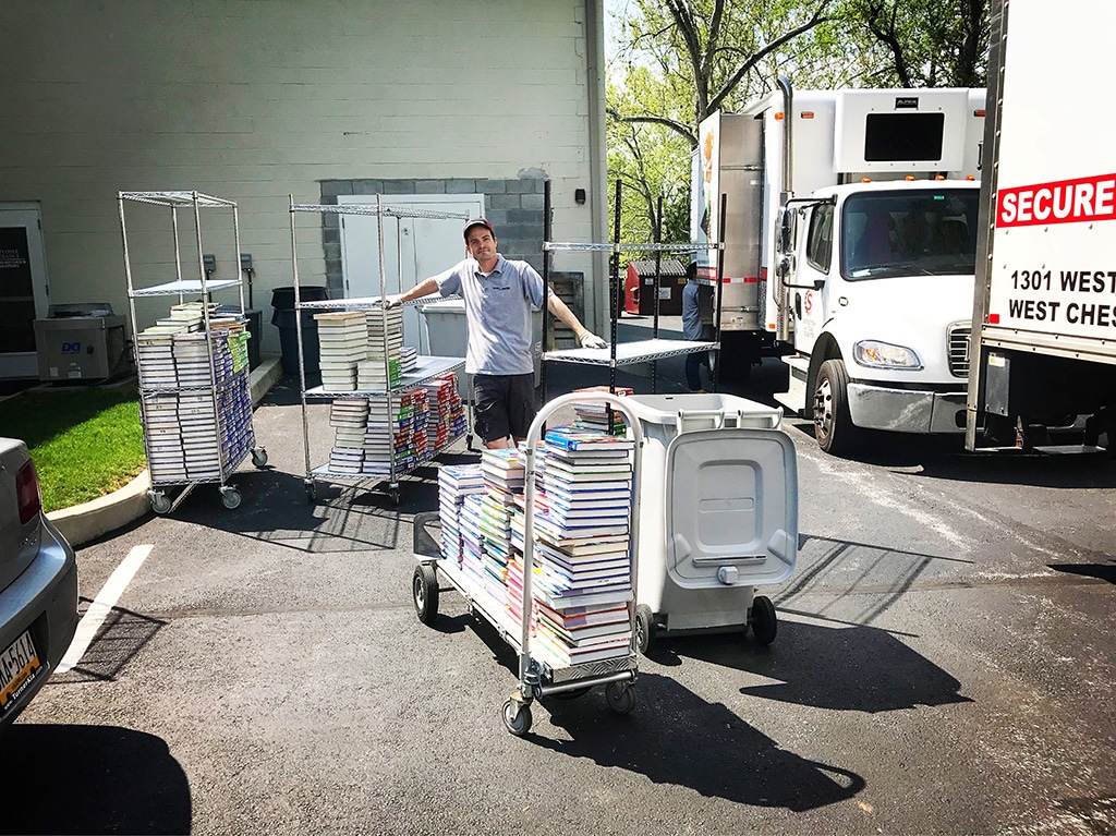 A man standing with multiple large carts of textbooks beside two Wiggins Shredding Trucks