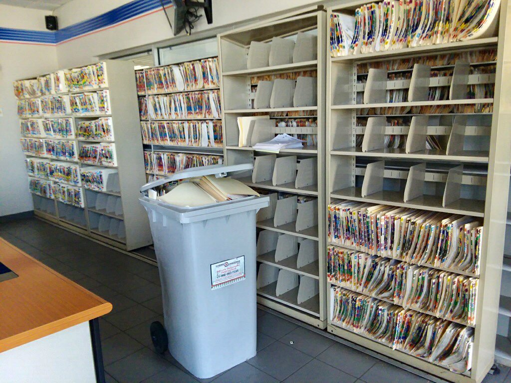 A secure shredding container in front of a wall of full shelves of documents