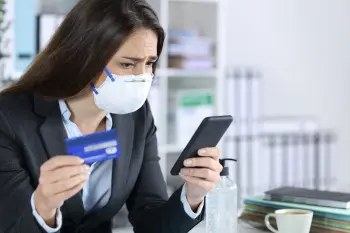 Woman with worried expression looking at her phone while holding a credit card and wearing a face mask