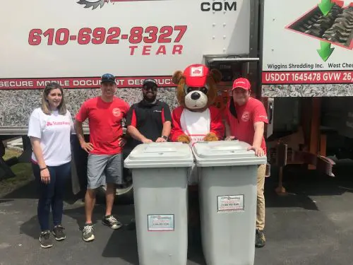 Four Individuals and a Bear Mascot stand with two secure collection containers in front of the Wiggins Shredding Truck