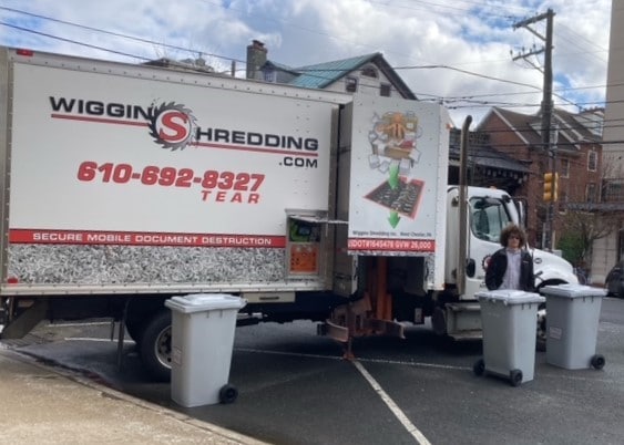 Large white shredding truck with Wiggins Shredding logo and a person standing in front of the truck with three large gray mobile shredding collection containers