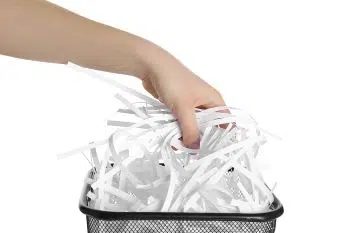 A person's hand grabbing long strips of white shredded paper out of a black mesh garbage can.