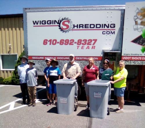 Four adults and four children standing at the side of a Wiggins shredding truck with two large gray shredding containers
