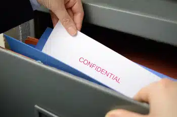 Hand pulling paper with the word, "Confidential" printed in red on it out of a hanging folder in a file cabinet