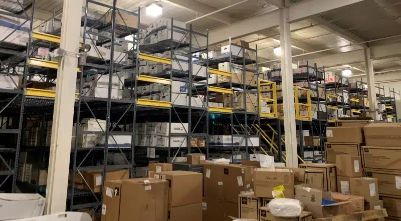 An interior photo of a warehouse. In the background, tall racking is filled with boxes, while in the foreground, more boxes are piled on the floor.