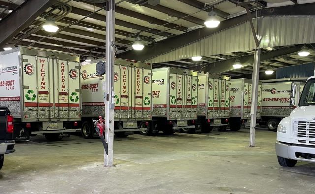 A photo showing a rear view of the fleet of Wiggins Shredding trucks lined up inside a warehouse truck bay area.
