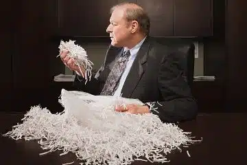 A man in a suit is sitting at a desk in a dimly-lit office and is holding a handful of shredded paper from the bag of shredded paper on the desk.