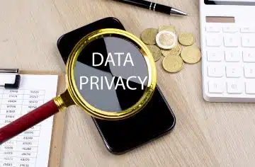 A magnifying glass is showing the words "Data Privacy" as it's being held over a mobile phone that's sitting on a desk surface, next to a clipboard, calculator and coins.