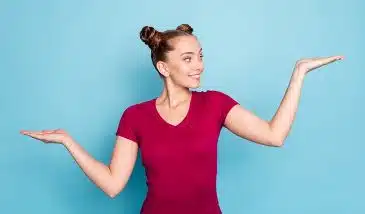 A smiling woman in a red t-shirt is holding her bent arms out to the sides with her palms facing upwards. One hand is higher than the other, indicating an unequal comparison between two things.